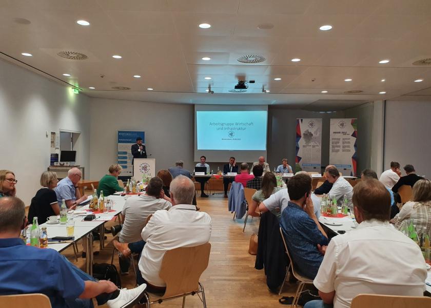 Ein Mann hält einen Vortrag, Menschen sitzen an Tischen und hören zu, im Hintergrund PowerPoint mit Arbeitsgruppe Wirtschaft und Infrastruktur