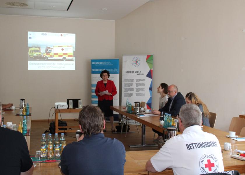 Ein Mann hält einen Vortrag, Menschen sitzen an Tischen und hören zu, im Hintergrund PowerPoint mit grenzüberschreiteneder Rettungsdienst im BY/CZ-Grenzgebiet