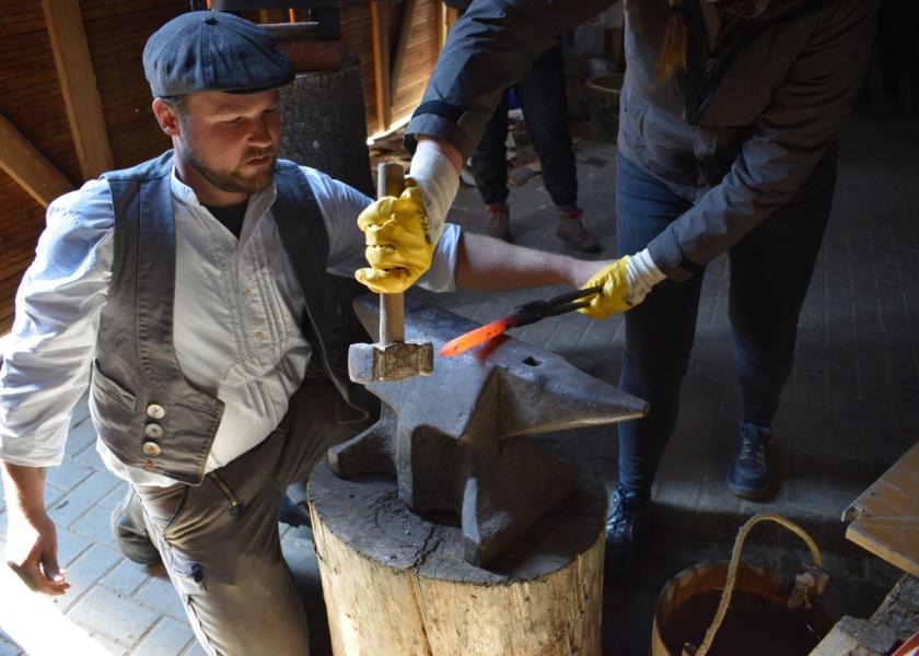 Ein Mann hilft einer Person mit einem Hammer in der Hand (Körper ohne Gesicht sichtbar) beim Schmieden