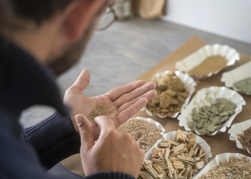 acht verschiedene Baustoffe in einem Behälter, ein Mann mit Rücken zur Kamera hat einen Baustoff in der linken Hand und fasst den Baustoff mit der rechten Hand an