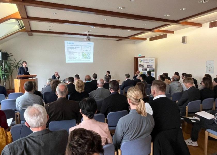 Eine Person hält einen Vortrag, Personen sitzen in einer Reihe und hören zu, im Hintergrund ist eine PowerPoint sichtbar