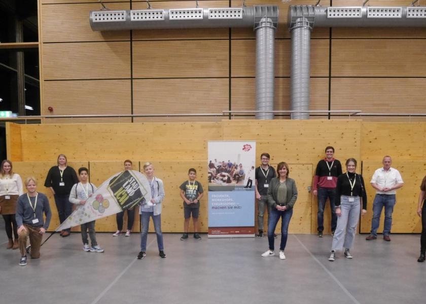 Insgesamt 13 Erwachsene und Kinder stehen vor einer Holzwand, in der Mitte ist ein RollUp zu Projekte, Workshops, Exkursionen - machen Sie mit!