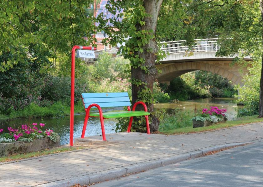 Bunte Mitfahrbank (Lehne: blau, Sitzfläche grün, Stelzen rot) sowie ein Schild mit dem Zielort
