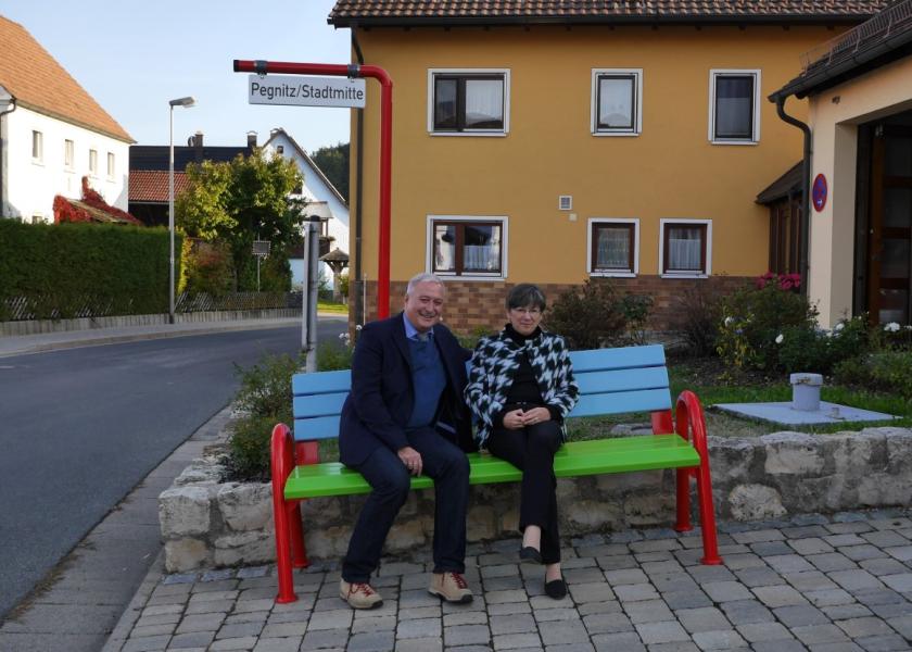 Eine Frau und ein Mann sitzen auf einer Mitfahrerbank, auf einem Schild ist der Zielort genannt: Pegnitz/Stadtmitte; im Hintergrund: Häuser