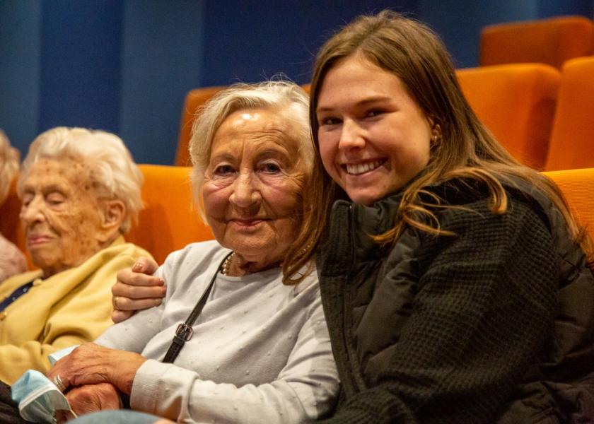 Eine Seniorin und ein junges Mädchen sitzen im Kino und lachen in die Kamera; im Hintergrund: weitere Seniorin sichtbar
