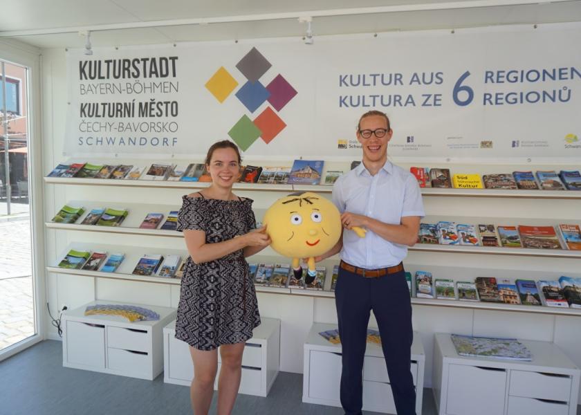 Eine junge Frau und ein junger Mann stehen im Kulturstadt-Pavillon mit einem Kuscheltier als Maskottchen in der Hand, im Hintergrund sind Broschüren ausgelegt