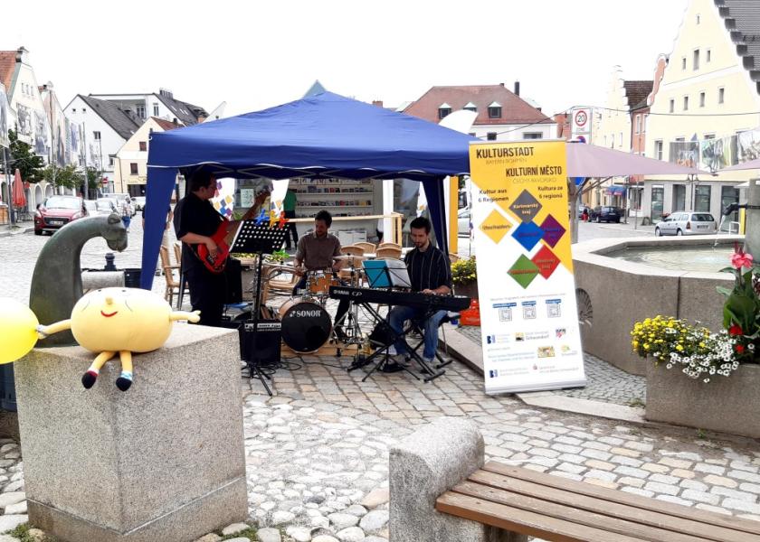 Eine Band mit Gitarre, Schlagzeug und ePiano spielen unter einem Pavillon auf einem Markplatz, im Vordergrund ist ein Kuscheltier als Maskottchen zu sehen, im Hintergrund der Kulturstadt-Pavillon