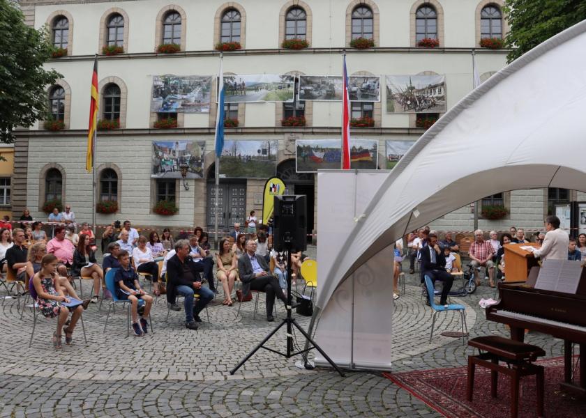 Menschen sitzen im Feien vor einem historischen Gebäude auf Stühlen und hren zu, rechts ist teilweise ein Pavillon mit einem Klavier zu sehen, eine Person steht am Rednerpult und hält eine Rede