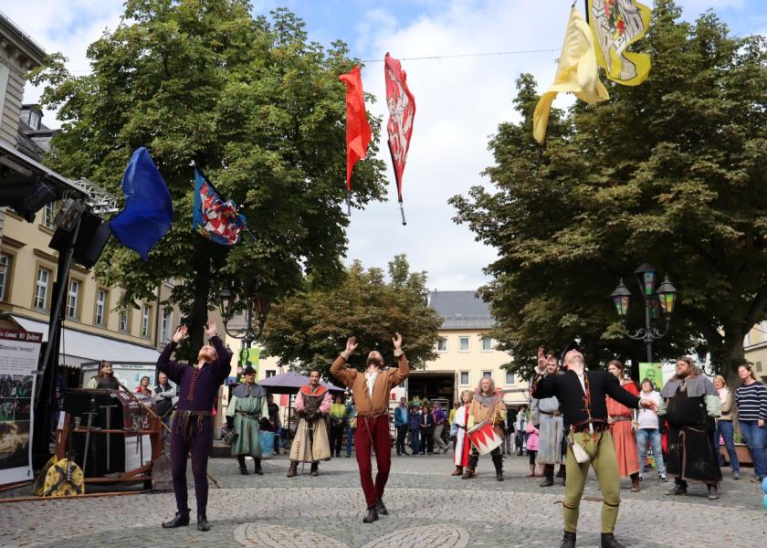 Drei Männer in mittelalterlicher Kleidung machen eine Vorführung und schmeißen ihre traditionellen Fahnen in die Luft, im Hintergrund sind weitere Personen in traditioneller Kleidung sowie Zuschauer 
