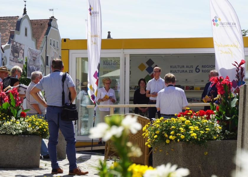 Kulturstadt-Pavillon auf einem Marktplatz inkl. Fahnen, eine Person stellt etwas vor, Personen außen rum hören aktiv zu und halten eine Broschüre in der Hand