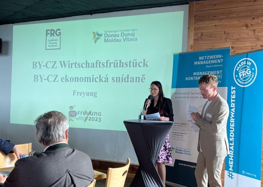Eine Frau hält einen Vortrag, daneben steht ein Mann, im Hintergrund: Leinwand mit BY-CZ Wirtschaftsfrühstück