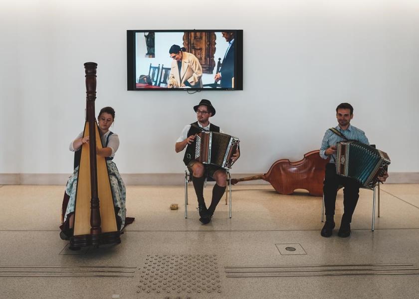drei Musiker in Lederhose bzw. Dirndl spielen Volksmusik, im Hintergrund sieht man einen kleinen Bildschirm, in dem eine Frau sich gerade in ein Buch einträgt