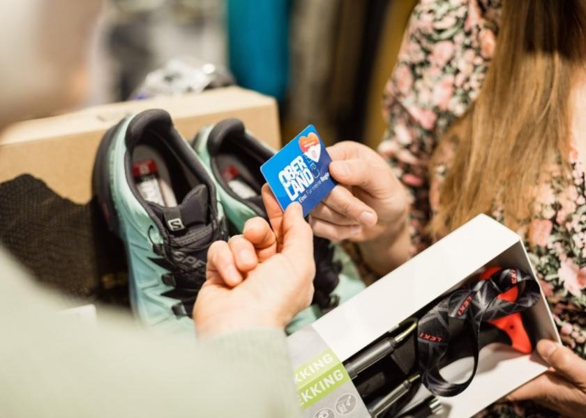 Auf dem Bild sind Wanderschuhe und Wanderstöcken in einem Outdoorladen sichtbar, zwei Personen halten die OBERLANDCard über die Schuhe und Stöcke