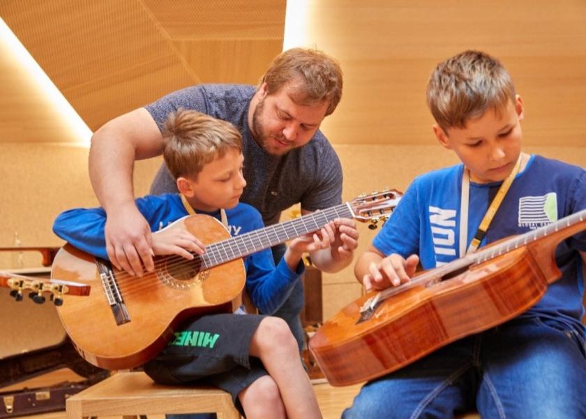 Zwei Kinder spielen Gitarre, ein Mann hilft einem Jungen