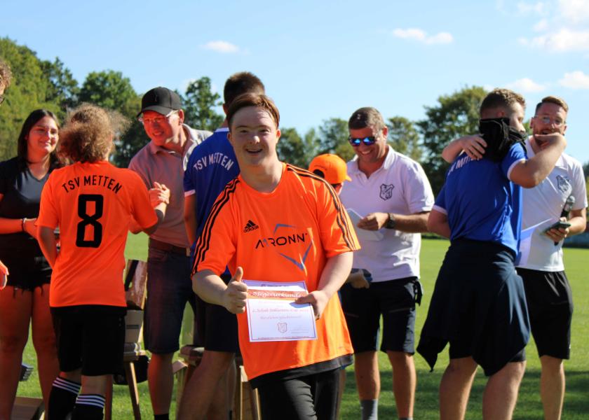im Vordergrund: Junge mit Downsyndrom, der Teilnehmerurkunde hält und Daumen hoch zeigt; im Hintergrund: weitere Turnierteilnehmer und Organisatoren des Inklusions-Fußballturniers