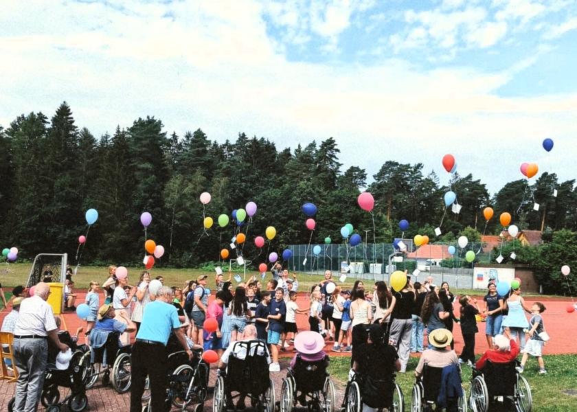 Gruppe aus Schülern und Senioren stehen auf Schulsportplatz im Kreis und lassen gemeinsam bunte Luftballons steigen