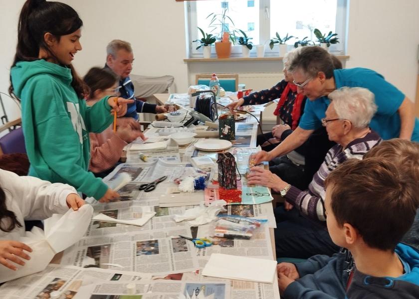 Gruppe aus Schülern und Senioren sitzen am Tisch, der mit Zeitungspapier ausgelegt ist, und basteln.
