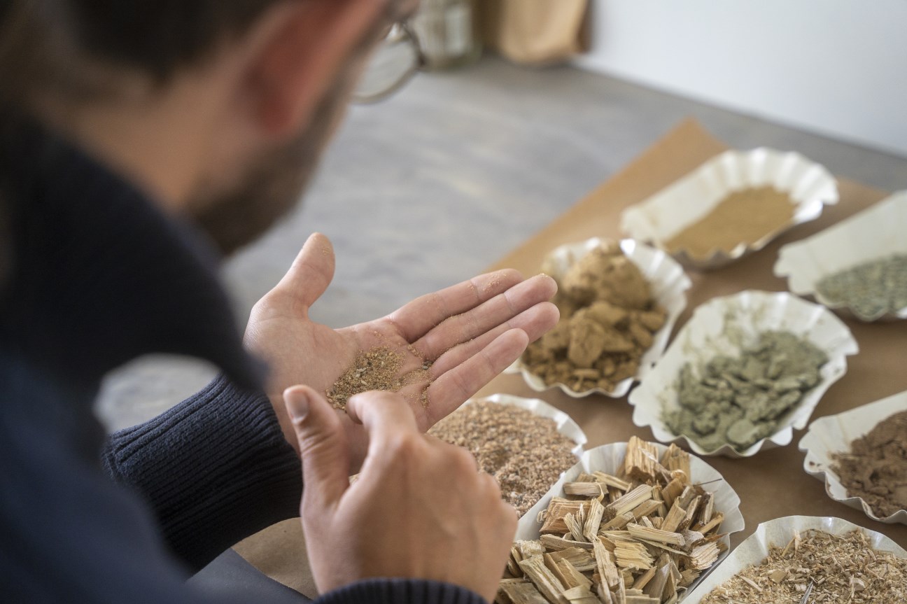 acht verschiedene Baustoffe in einem Behälter, ein Mann mit Rücken zur Kamera hat einen Baustoff in der linken Hand und fasst den Baustoff mit der rechten Hand an