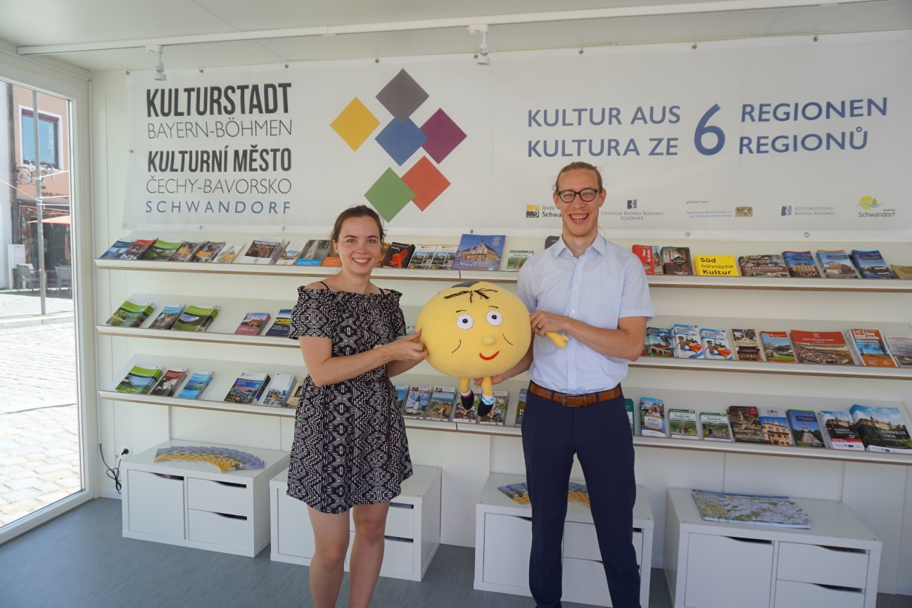 Eine junge Frau und ein junger Mann stehen im Kulturstadt-Pavillon mit einem Kuscheltier als Maskottchen in der Hand, im Hintergrund sind Broschüren ausgelegt