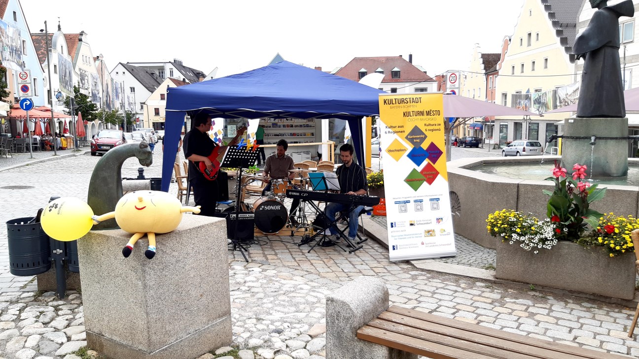 Eine Band mit Gitarre, Schlagzeug und ePiano spielen unter einem Pavillon auf einem Markplatz, im Vordergrund ist ein Kuscheltier als Maskottchen zu sehen, im Hintergrund der Kulturstadt-Pavillon