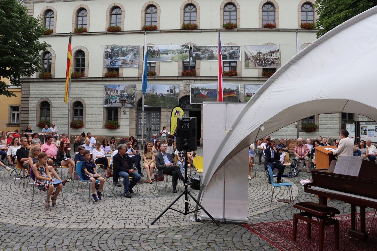 Menschen sitzen im Feien vor einem historischen Gebäude auf Stühlen und hren zu, rechts ist teilweise ein Pavillon mit einem Klavier zu sehen, eine Person steht am Rednerpult und hält eine Rede