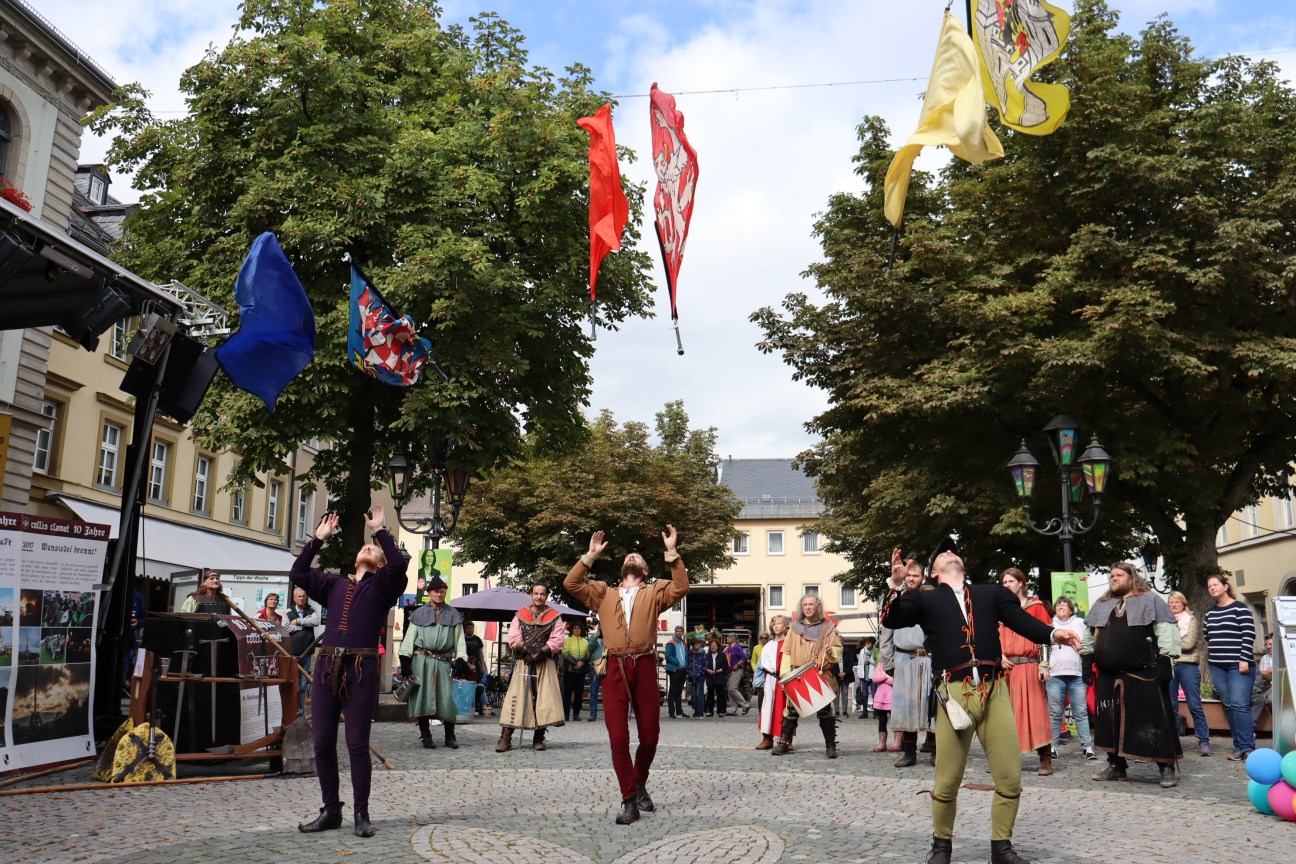 Drei Männer in mittelalterlicher Kleidung machen eine Vorführung und schmeißen ihre traditionellen Fahnen in die Luft, im Hintergrund sind weitere Personen in traditioneller Kleidung sowie Zuschauer 