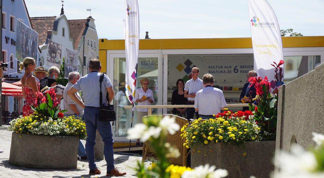 Kulturstadt-Pavillon auf einem Marktplatz inkl. Fahnen, eine Person stellt etwas vor, Personen außen rum hören aktiv zu und halten eine Broschüre in der Hand