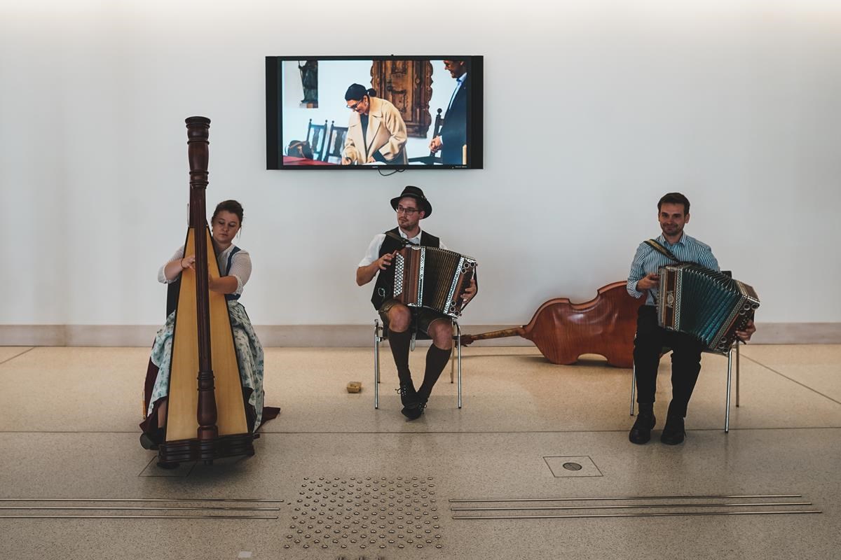 drei Musiker in Lederhose bzw. Dirndl spielen Volksmusik, im Hintergrund sieht man einen kleinen Bildschirm, in dem eine Frau sich gerade in ein Buch einträgt