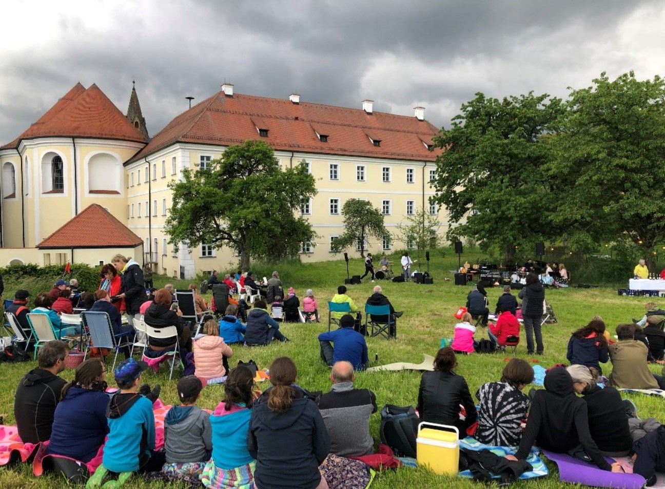 Menschen sitzen auf einer Wiese mit und ohne Stühle und hören einer Band zu, im Hintergrund ist das Kloster Frauenzell 