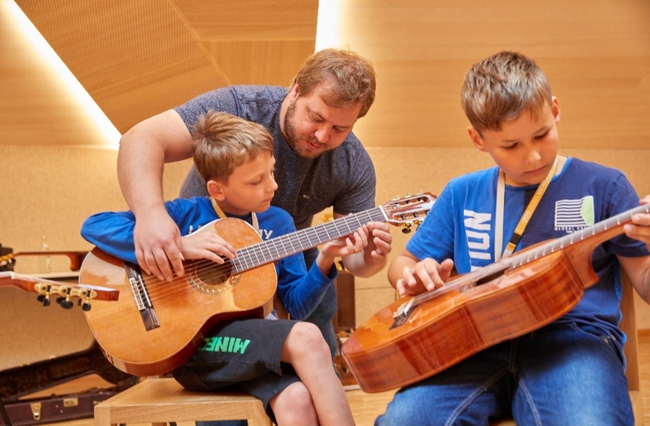 Zwei Kinder spielen Gitarre, ein Mann hilft einem Jungen 