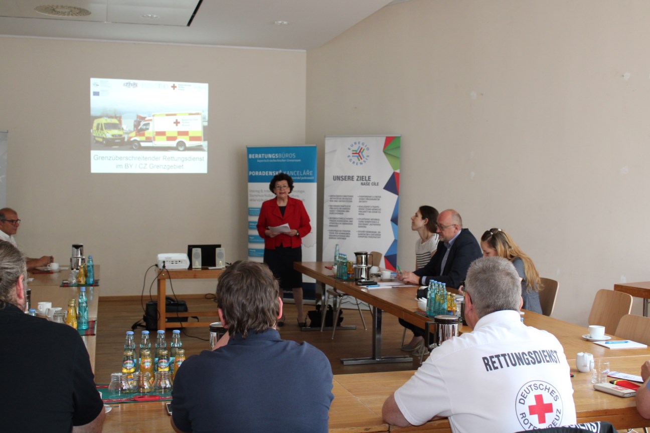 Ein Mann hält einen Vortrag, Menschen sitzen an Tischen und hören zu, im Hintergrund PowerPoint mit grenzüberschreiteneder Rettungsdienst im BY/CZ-Grenzgebiet