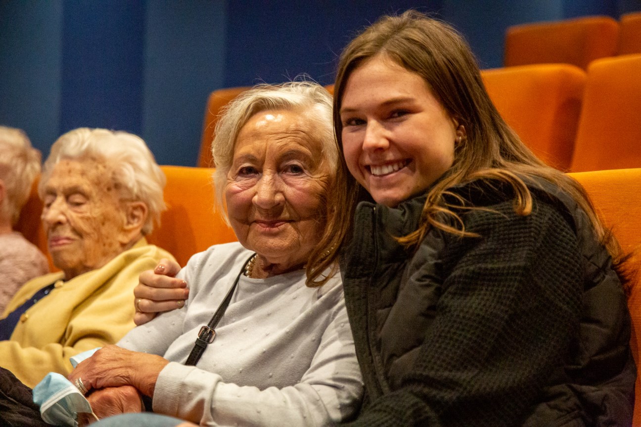 Eine Seniorin und ein  junges Mädchen sitzen im Kino und lachen in die Kamera; im Hintergrund: weitere Seniorin sichtbar