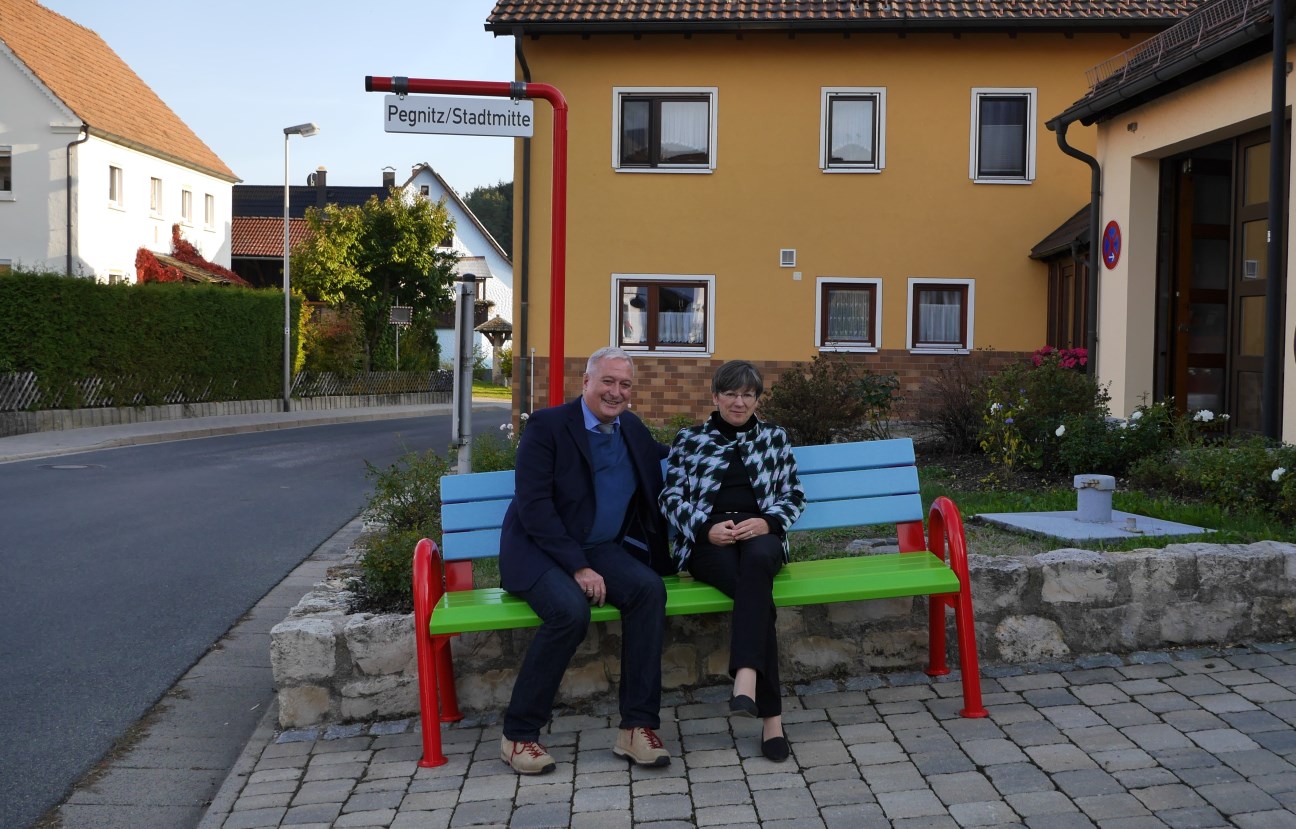 Eine Frau und ein Mann sitzen auf einer Mitfahrerbank, auf einem Schild ist der Zielort genannt: Pegnitz/Stadtmitte; im Hintergrund: Häuser