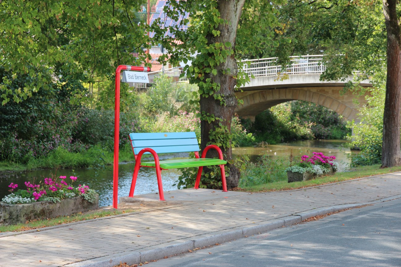 Bunte Mitfahrbank (Lehne: blau, Sitzfläche grün, Stelzen rot) sowie ein Schild mit dem Zielort &quot;Bad Berneck&quot;,; im Hintergrund ist ein Fluss und eine Brücke sichtbar